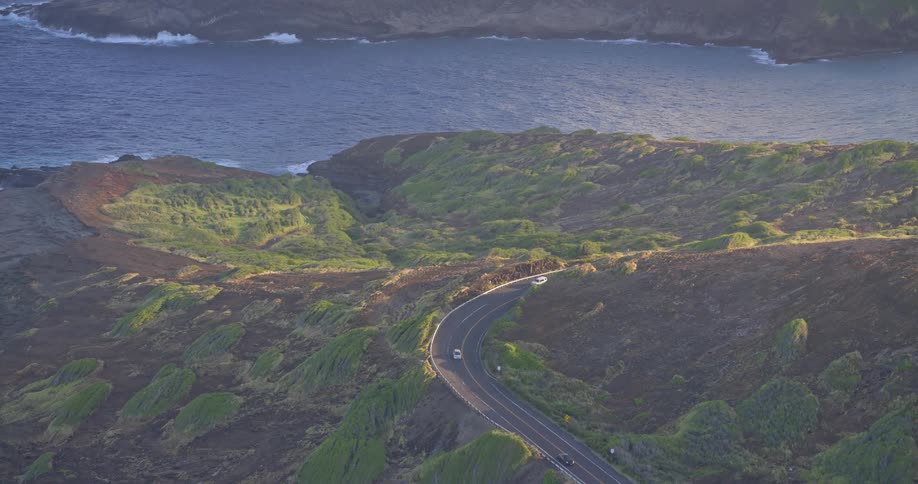 A scenic high-angle view shows cars driving along a winding coastal highway in Hawaii during golden hour