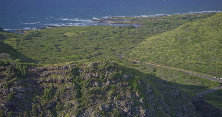 An aerial view slowly rises over a rugged green cliff to reveal a beautiful coastline with a seaside highway and the deep blue ocean