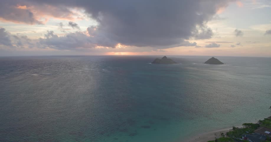 An aerial pull-back shot reveals a tropical coastline with houses and palm trees overlooking a vast ocean with two distant islands at sunrise