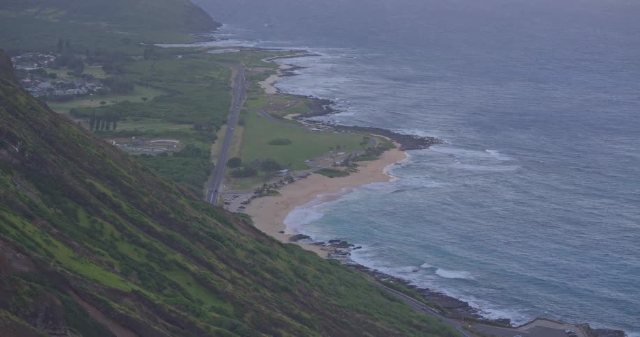A stunning high-angle, slow zoom-out shot reveals the dramatic coastline of Oahu, Hawaii, with cars driving on a coastal road next to a sandy beach at dusk
