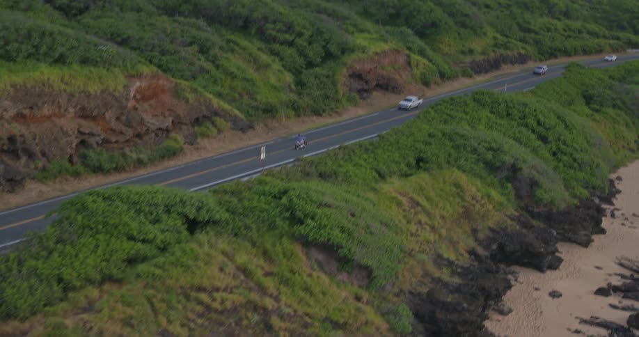 An aerial view captures a lone motorcyclist cruising down a winding coastal highway flanked by lush green hills and a sandy beach