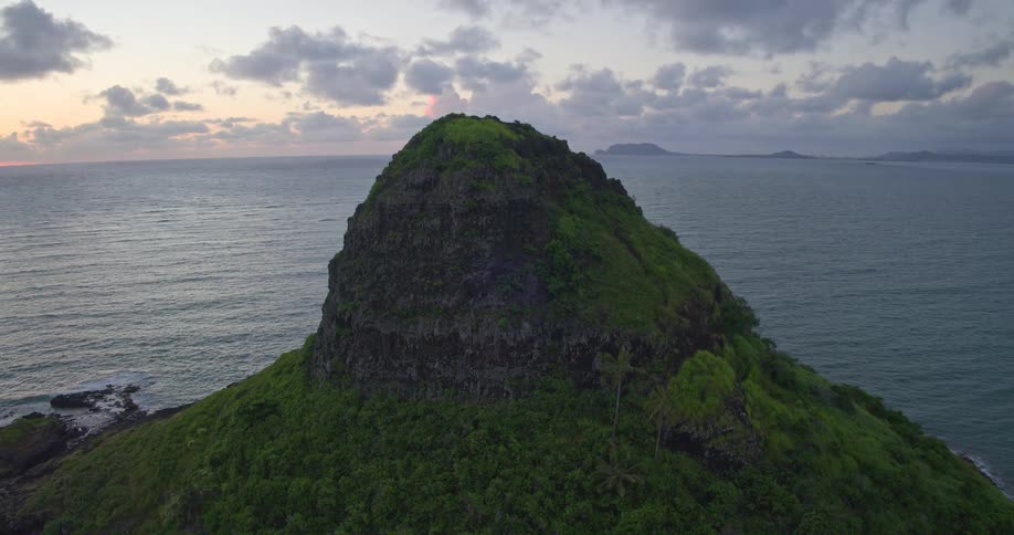 An expansive aerial shot slowly pulls back from a dramatic, lush green tropical island during a beautiful sunrise over the ocean