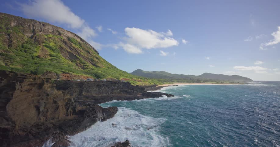 A stunning aerial view reveals powerful ocean waves crashing against the rugged volcanic cliffs and a blowhole on a sunny day along a lush tropical coastline