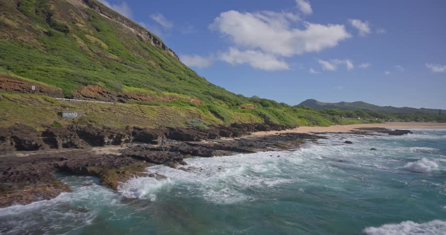 A slow aerial view follows the rugged, rocky coastline of a tropical island, where turquoise waves crash against the shore below a lush green mountain