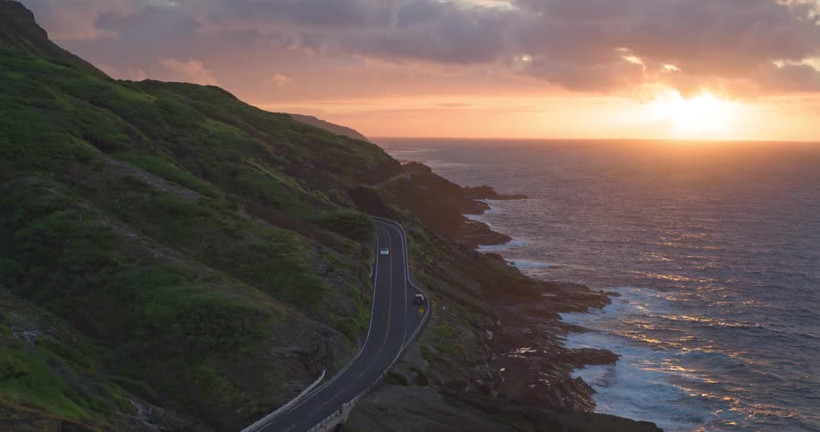 A stunning high-angle view captures cars driving on a winding coastal highway alongside a lush green mountain during a beautiful sunset over the ocean