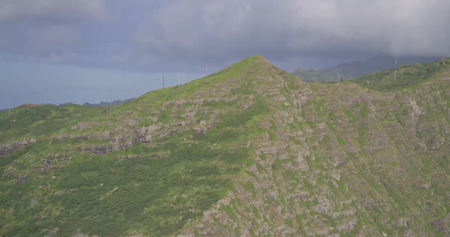 Sweeping aerial pan across a lush green mountain range revealing the distant Hawaiian coastline under a dramatic cloudy sky