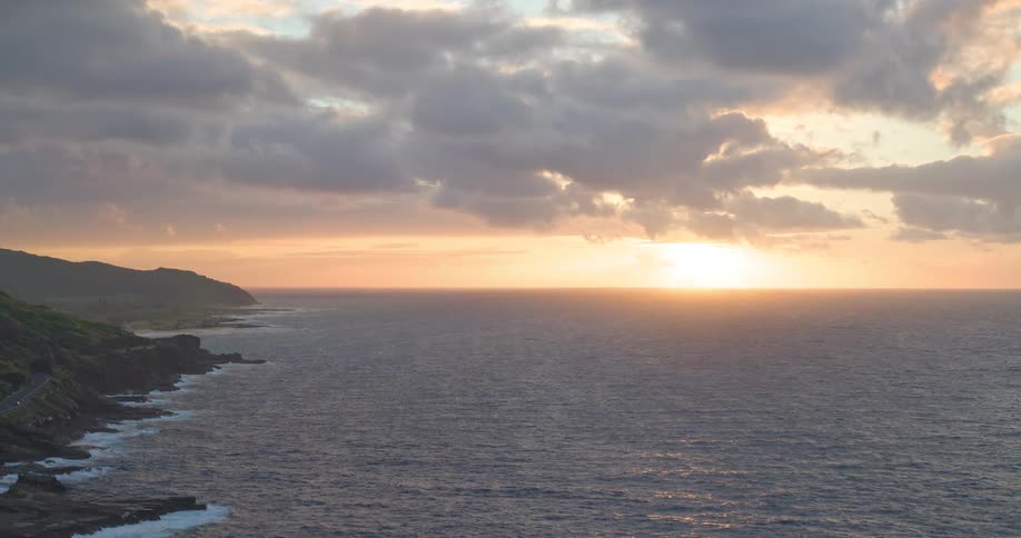 A serene sunrise casts a warm glow over a calm ocean and a rocky, green coastline under a cloudy sky