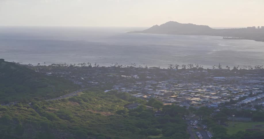 An aerial view captures the lush Hawaiian coast, with a city by the ocean and Diamond Head silhouetted in the hazy sunlight