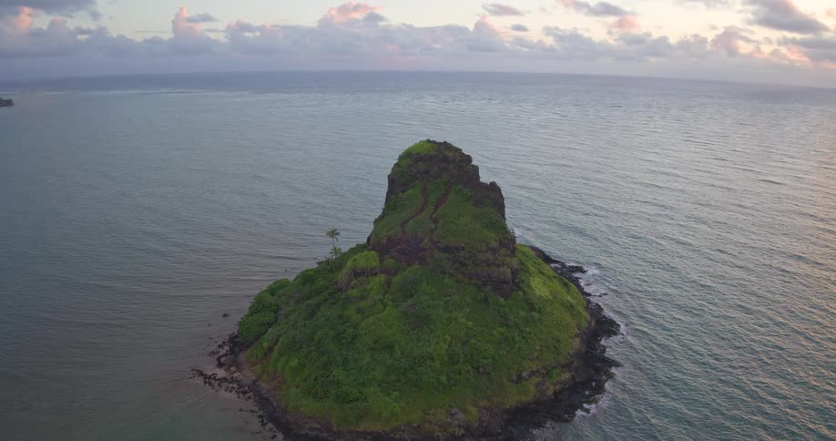 An aerial ascent reveals the beauty of Mokolii (Chinaman's Hat) island in Hawaii, set against a backdrop of coastal mountains and a colorful sunrise