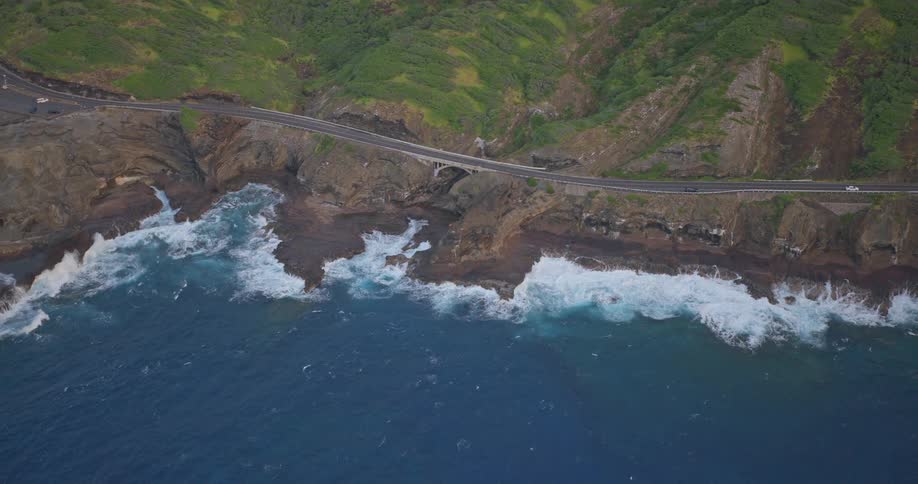 Stunning aerial view pulls back to reveal a scenic highway winding along a rugged, green coastline as powerful blue waves crash against the rocky shore