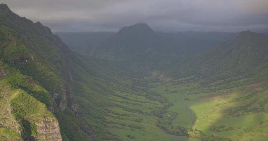 An epic aerial view pulls back to reveal a lush mountain valley under a dramatic, cloudy sky