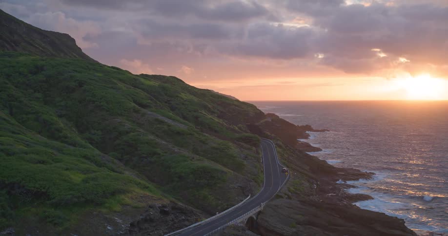 Stunning aerial view of a scenic coastal highway winding along lush green cliffs as the sun sets over the ocean