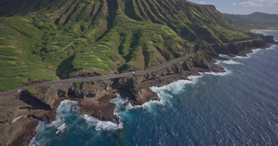 A stunning aerial shot tilts down over a scenic coastal highway where cars drive along the dramatic, rocky shoreline at the base of a lush green mountain