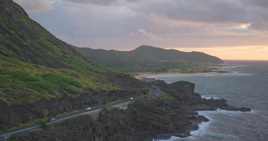 Aerial pullback shot reveals cars driving on a winding coastal highway along the stunning Hawaiian coastline at sunrise