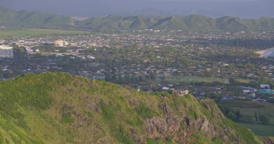 An aerial view flies towards a scenic mountain lookout point where hikers enjoy panoramic views of the Hawaiian coastline and a distant town