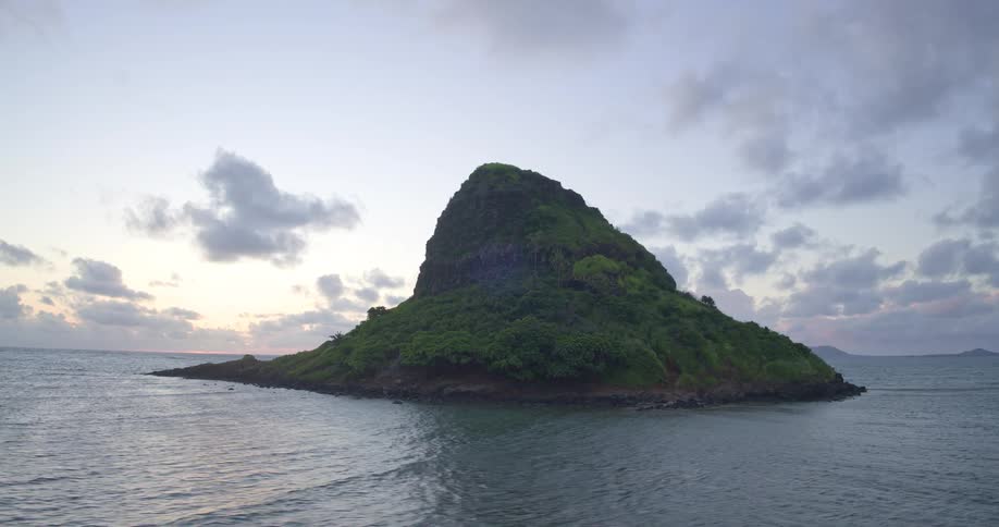 Cinematic aerial view slowly orbits the lush, iconic Mokolii island, known as Chinaman's Hat, surrounded by calm ocean waters at sunrise off the coast of Oahu, Hawaii