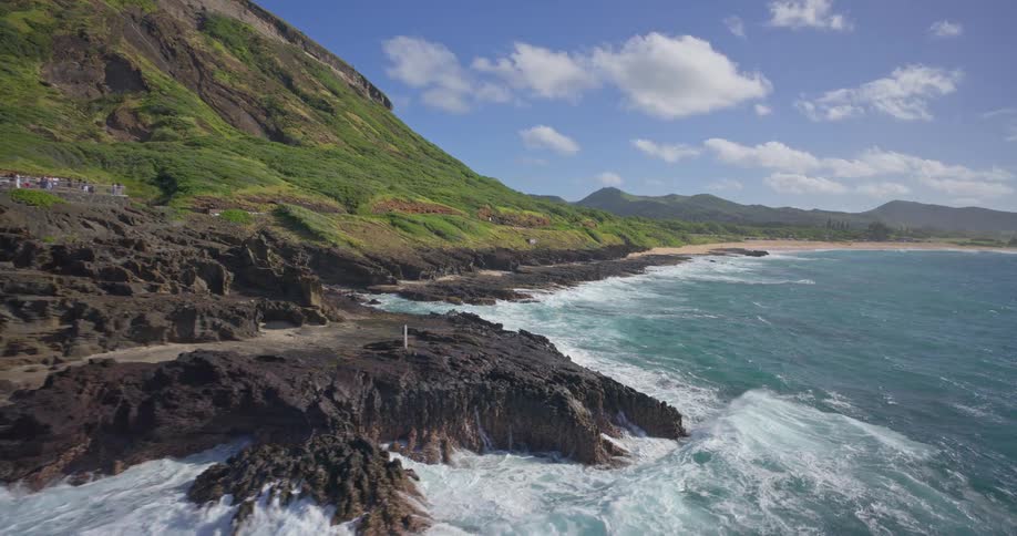 An aerial view slowly advances along the dramatic volcanic coastline of Oahu, Hawaii, where powerful waves crash against the black rocks