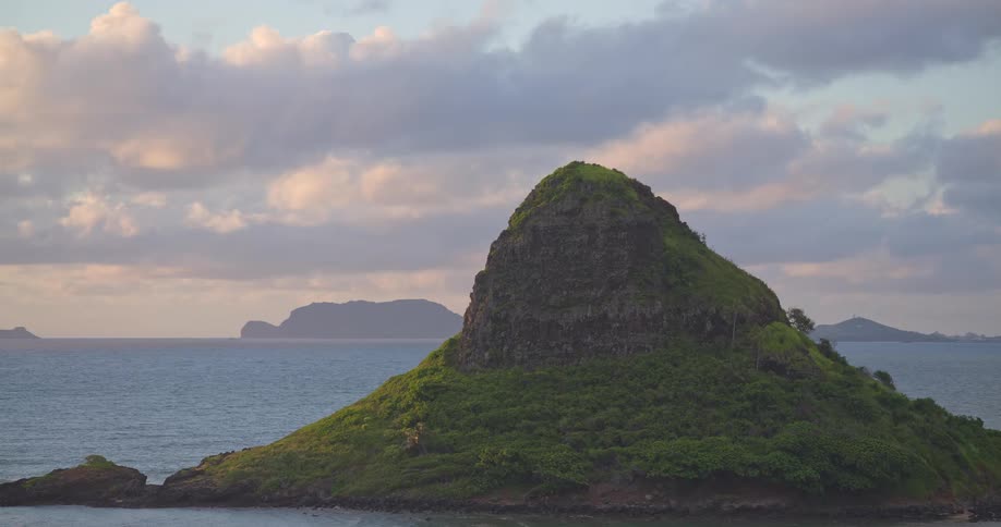 A cinematic pan reveals the iconic Mokolii islet, also known as Chinaman's Hat, off the coast of Oahu, Hawaii, during a peaceful sunrise