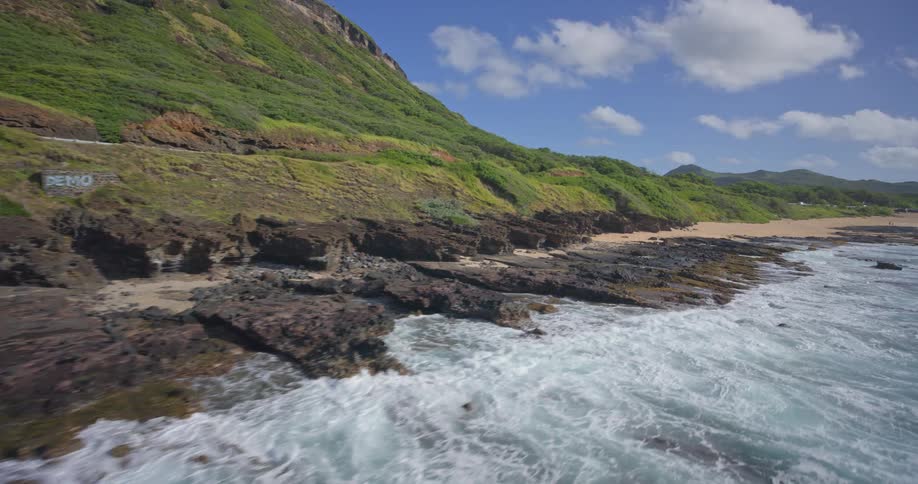 An aerial view tracking slowly along a rugged and lush green tropical coastline as turquoise waves crash against the rocky shore