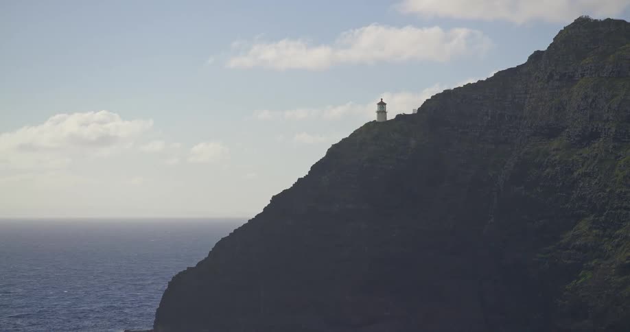 A solitary lighthouse stands watch over the vast blue ocean from its perch on a steep, verdant cliffside