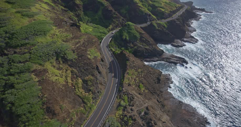 An incredible aerial view follows a winding coastal highway as it snakes between lush green mountainsides and the sparkling blue Pacific Ocean