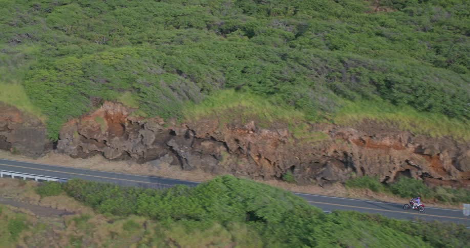 A lone rider on a motorcycle travels down a scenic coastal highway carved into lush green hillsides
