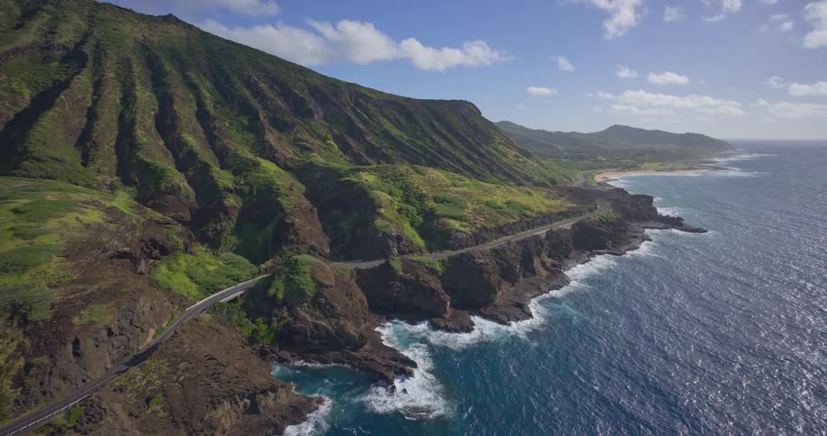Stunning aerial view pulling back from a scenic coastal highway nestled between lush green mountains and the deep blue Pacific Ocean