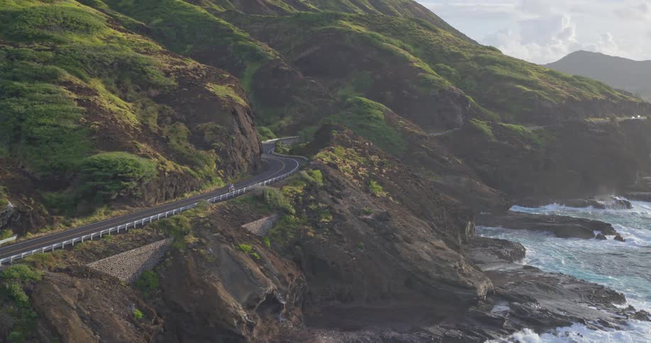 An aerial view shows a motorcycle driving along a scenic and winding coastal highway carved into the side of a lush, green mountain cliffside