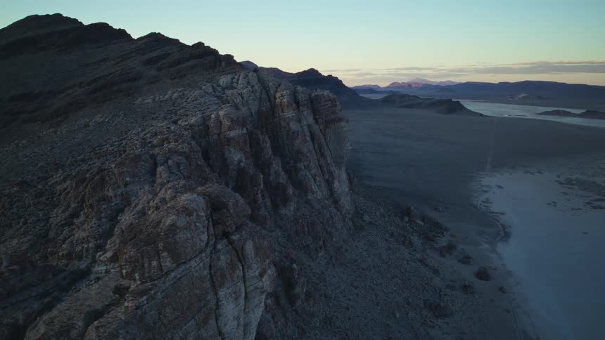 Slow aerial dolly shot along a dramatic, rocky mountain ridge overlooking a vast desert landscape at sunrise