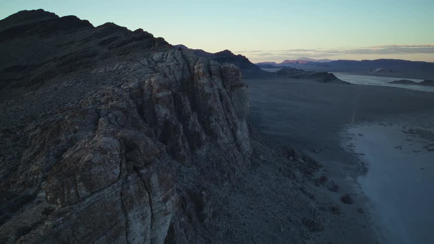 Slow aerial dolly shot along a dramatic, rocky mountain ridge overlooking a vast desert landscape at sunrise