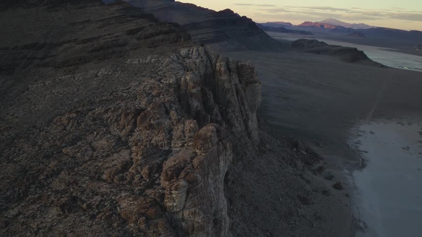 An aerial view reveals the vast, rocky landscape of a desert mountain range bordering a dry salt flat at sunrise