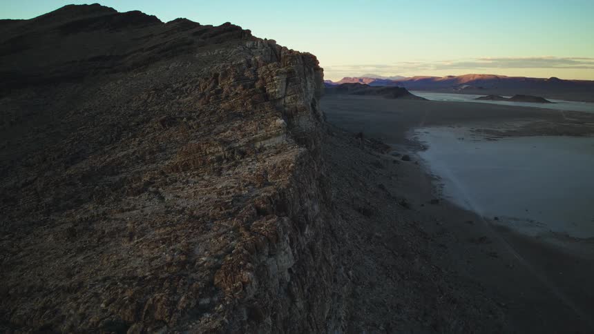 A static wide shot of a rugged cliff face overlooking a vast desert valley and dry lake bed at sunrise