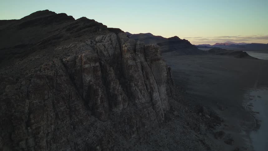 An epic aerial shot reveals a vast, rugged mountain range overlooking a desolate desert landscape at twilight