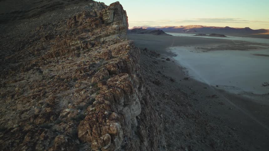 A slow aerial pan up a rocky cliff reveals a vast, dry desert lakebed at sunrise