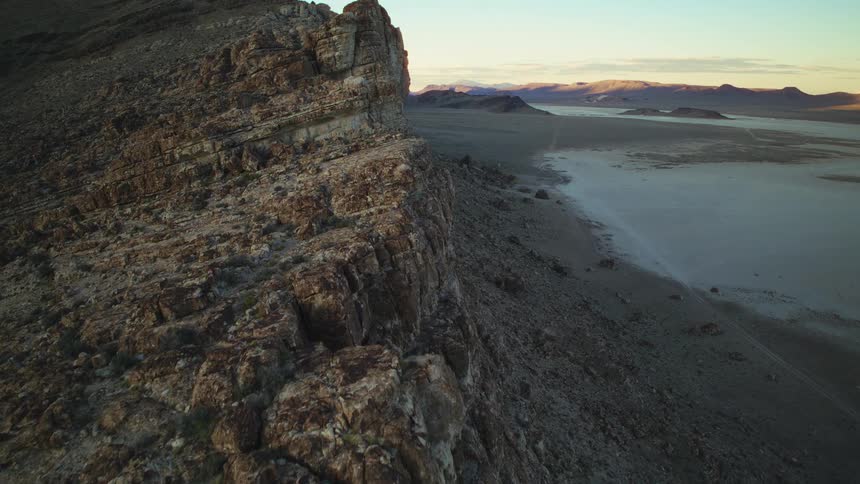 A slow aerial pan up a rocky cliff reveals a vast, dry desert lakebed at sunrise