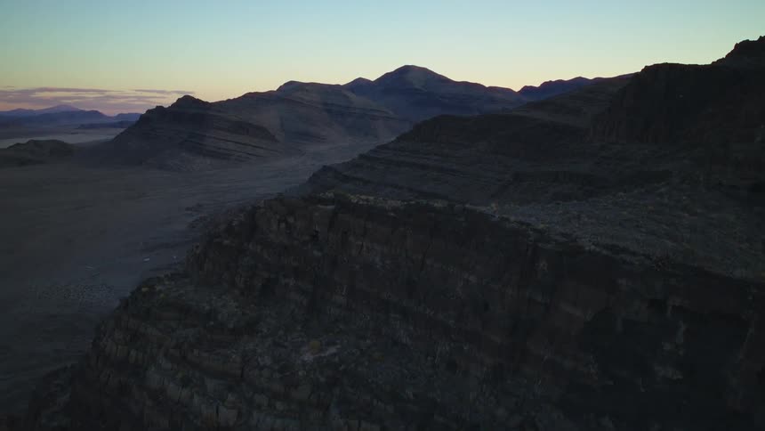 An aerial view ascends over a rugged, layered mountain ridge in a vast desert landscape at twilight