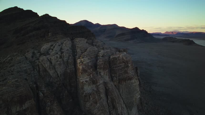 A slow aerial dolly shot pulls back from a dramatic desert cliff face, revealing the vast mountain landscape at dusk
