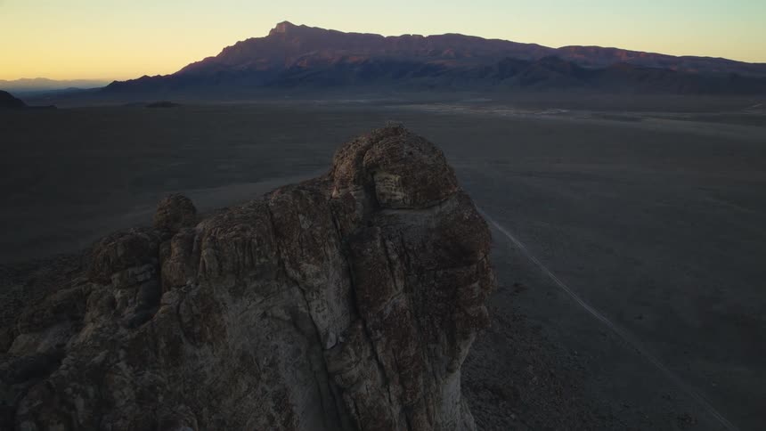 A cinematic aerial view slowly approaches a rugged cliff in a vast desert landscape as the sun rises over a distant mountain range