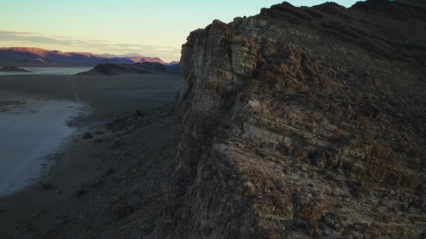 A slow and majestic aerial push-in shot along a rugged mountain cliff reveals a vast desert valley at golden hour