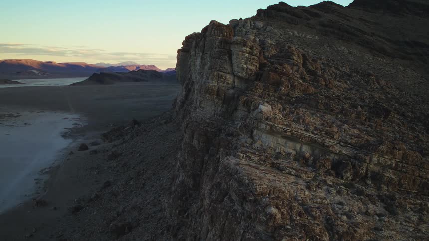 A slow and majestic aerial push-in shot along a rugged mountain cliff reveals a vast desert valley at golden hour