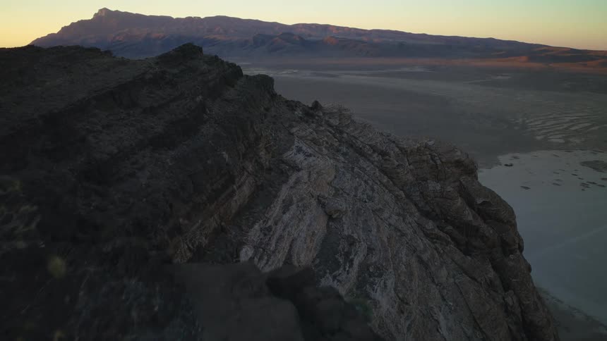 An aerial view slowly advances along a dark, rocky ridge overlooking a vast desert valley and distant mountains at sunrise