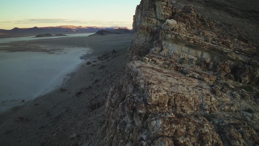 A slow cinematic aerial shot flies along the edge of a massive, rocky cliff overlooking a vast desert landscape at sunrise