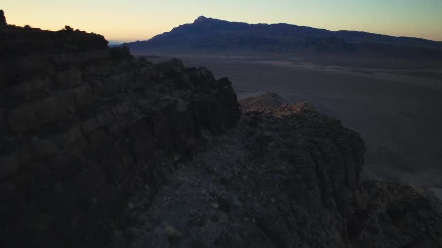 An epic aerial shot pushes over a shadowed mountain ridge at dusk, revealing a vast desert landscape below