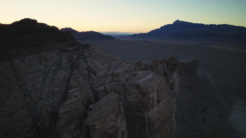 An aerial shot slowly pulls back along a craggy mountain ridge overlooking a vast desert landscape at twilight