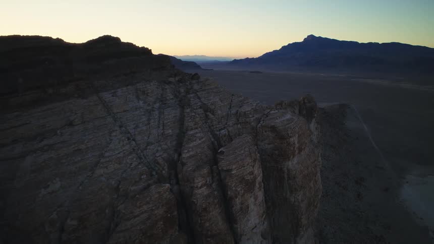 An aerial shot slowly pulls back along a craggy mountain ridge overlooking a vast desert landscape at twilight