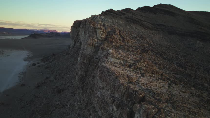 A majestic, stationary aerial shot captures the warm glow of sunset on a towering cliff overlooking a dry desert playa