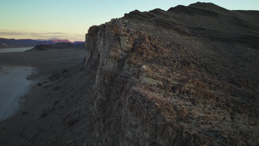 A majestic, stationary aerial shot captures the warm glow of sunset on a towering cliff overlooking a dry desert playa