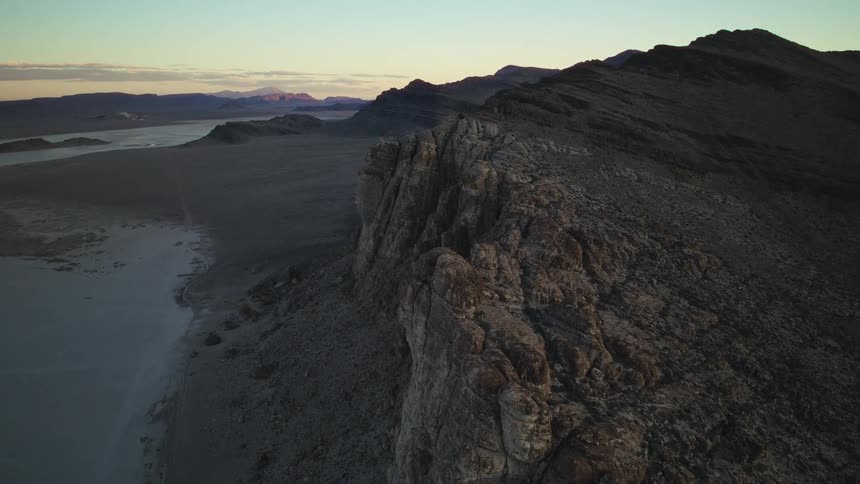 An aerial view shows a rugged mountain ridge overlooking a vast desert plain during a serene sunset