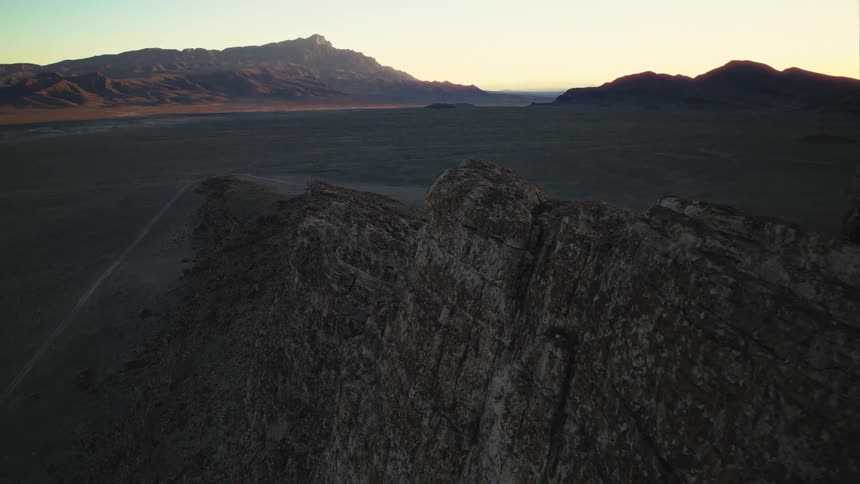Drone footage slowly reveals a majestic desert valley and distant mountains from behind a rocky ridge at sunrise