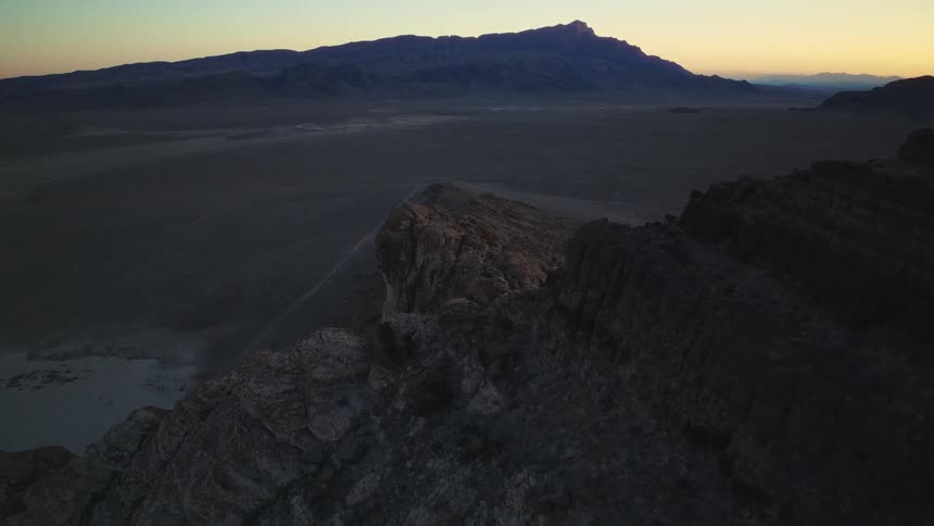 An aerial view slowly advances along a rugged mountain crest above a wide desert valley during twilight